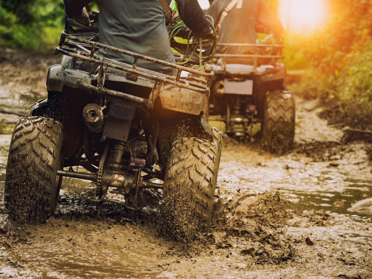 Two people riding ATVs through a muddy off-road trail on a sunny day.
