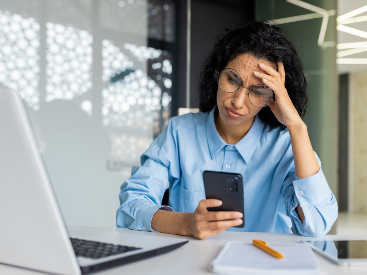 Business owner looking stressed while reading a Cyber alert on a smartphone beside a laptop