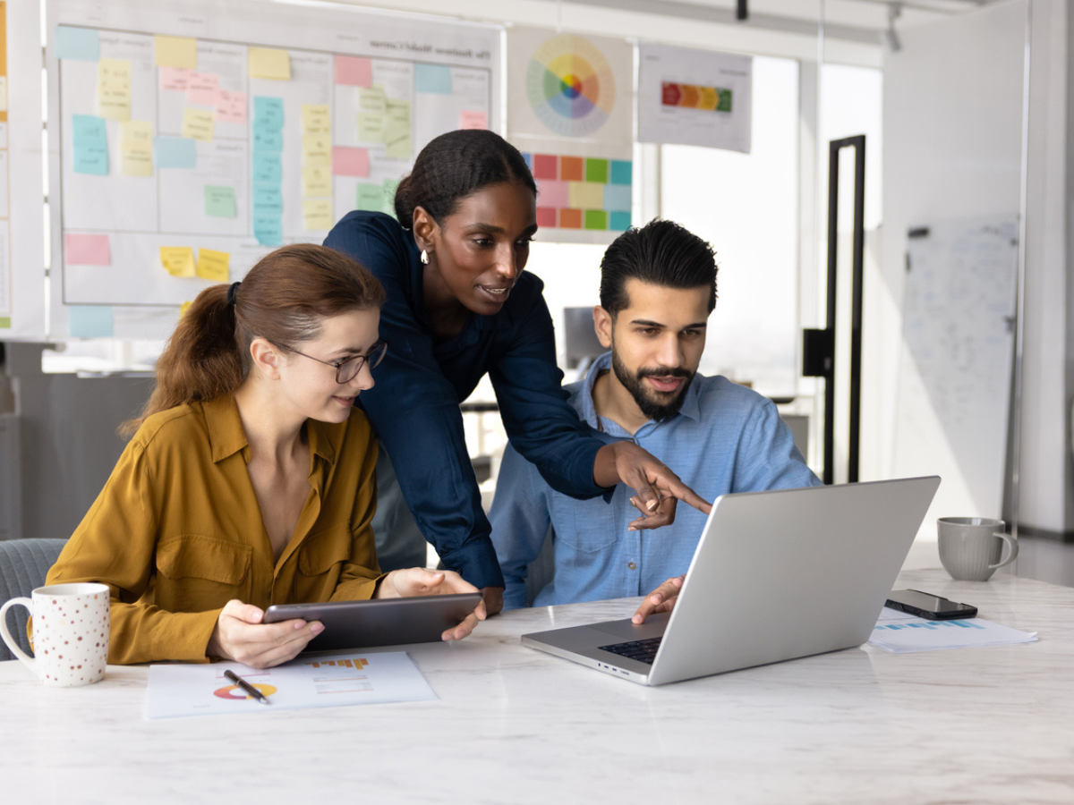 Three colleagues collaborating at a desk, reviewing information on a laptop and tablet in a modern office