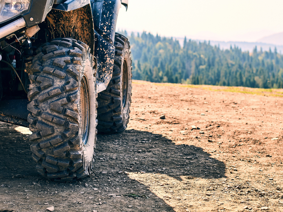 Close-up of deep-tread ATV tires on a dirt trail with a scenic mountain forest in the background.