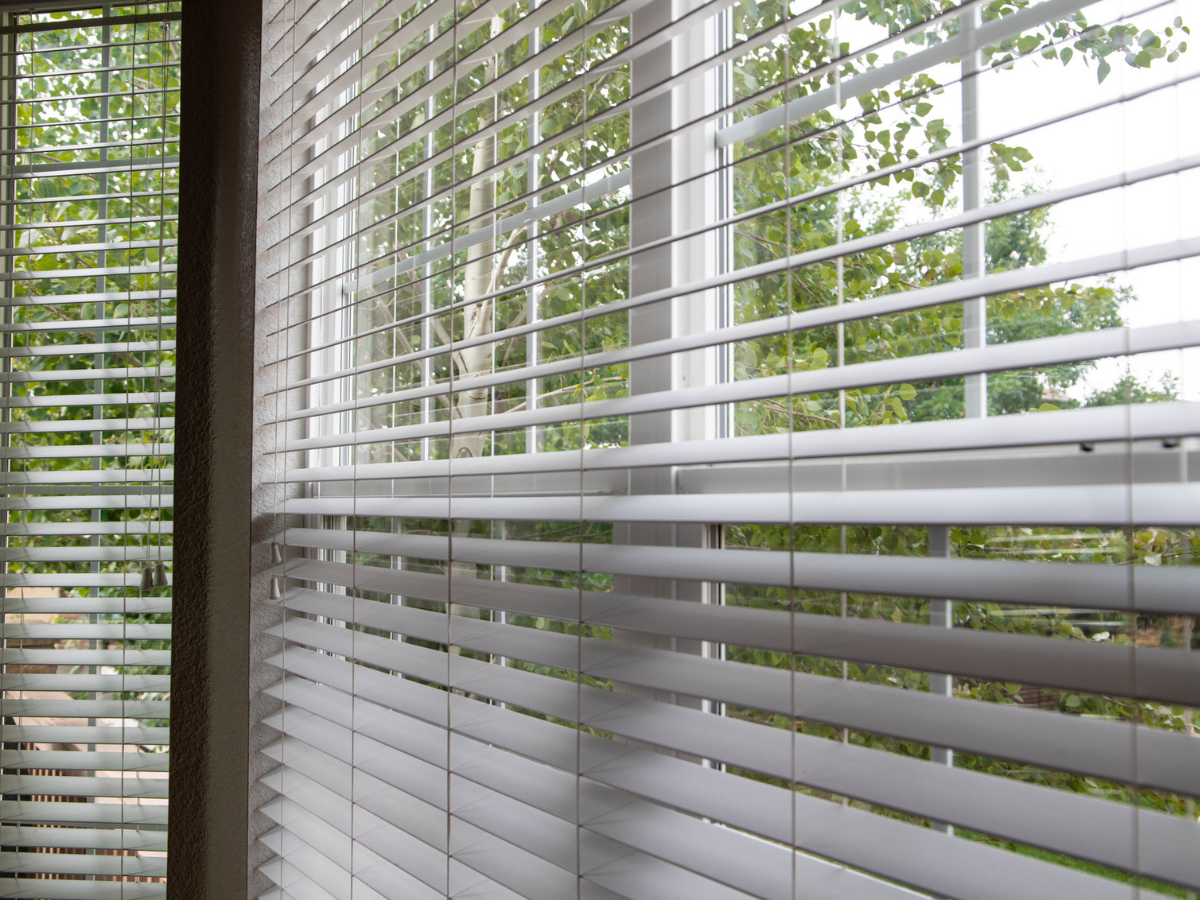 Partially closed window blinds inside a home with trees visible outside.