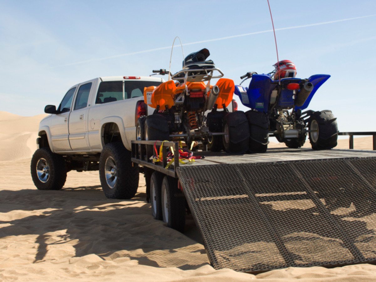 Two ATVs loaded on a flatbed trailer towed by a pickup truck in sand dunes