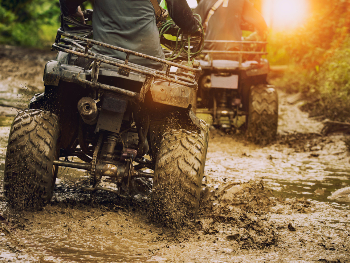 Two people riding ATVs through a muddy off-road trail on a sunny day.