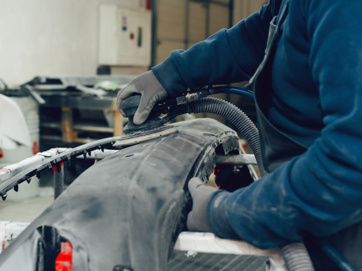 Mechanic sanding a car bumper with a power tool to prepare a restored vehicle in an auto shop.