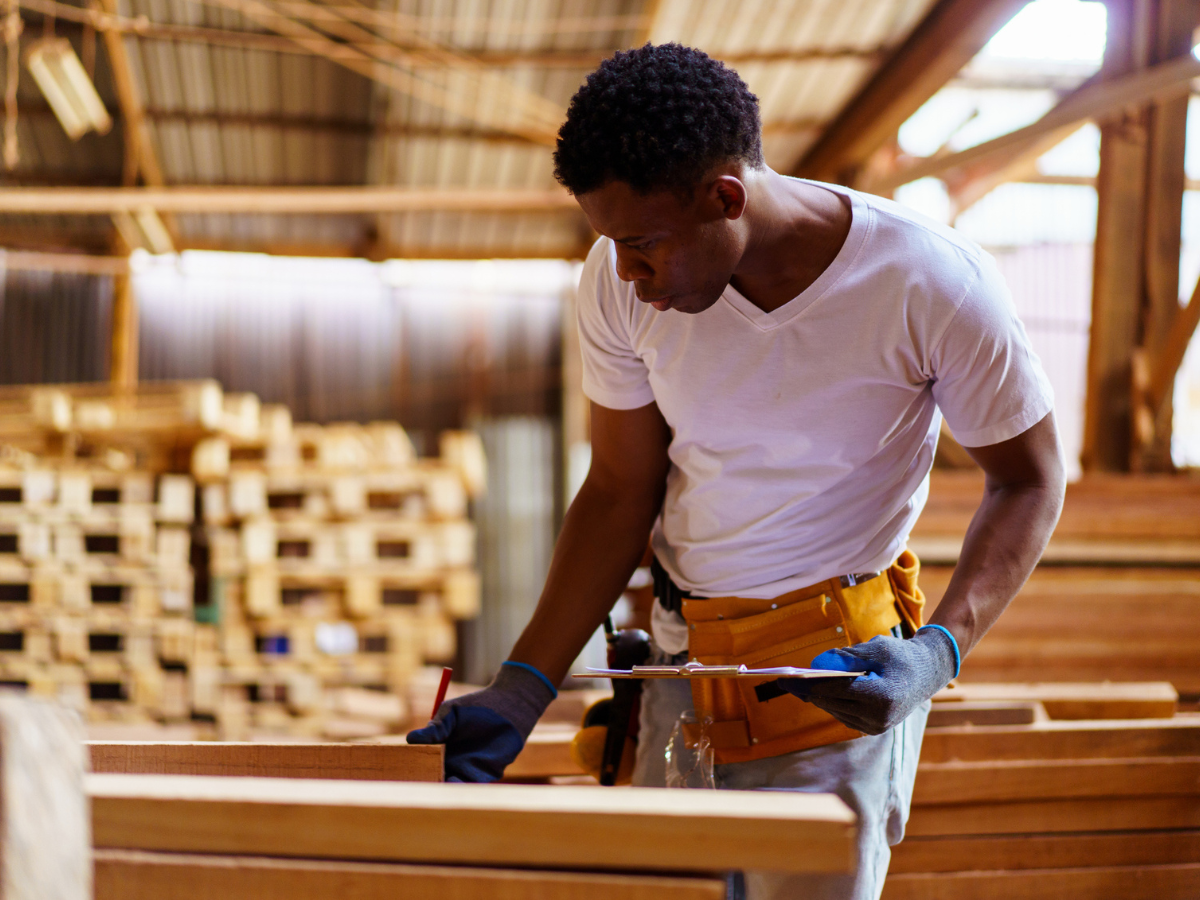 Carpenter wearing work gloves and a tool belt measures and marks wooden boards inside a workshop.
