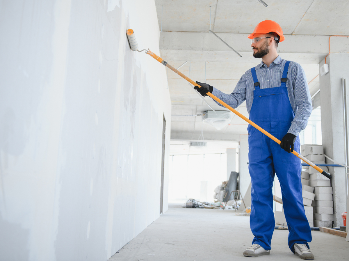 Painter wearing safety gear uses a roller to apply white paint to an interior wall on a construction site.