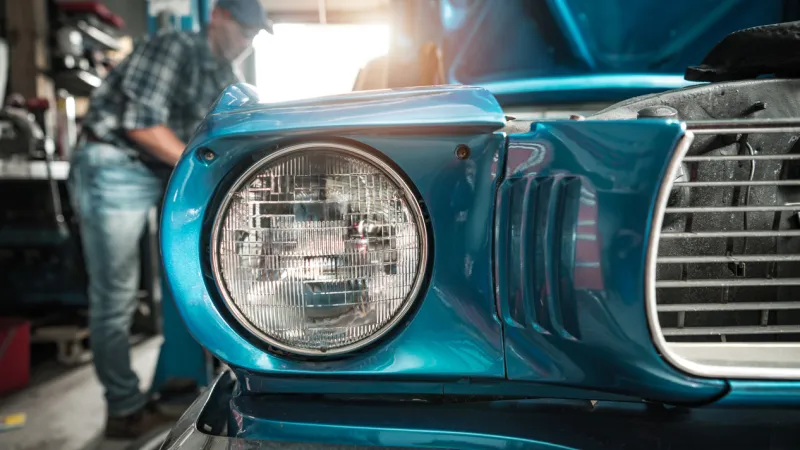 Close up of a restored vintage car headlight in a garage with a mechanic working in the background, representing classic vehicle restoration and insurance